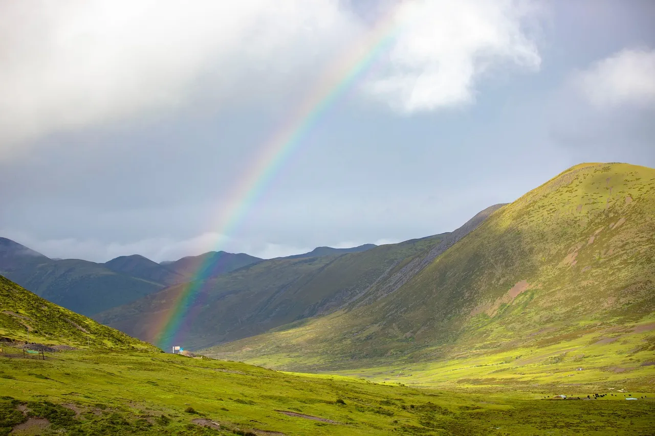 A vibrant rainbow arcs across a dramatic mountain valley with rolling green hills, scattered sheep, and cloudy skies
